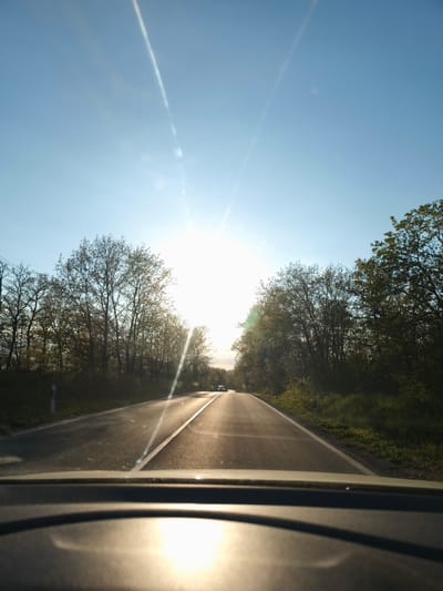 The image depicts a road in Zdravets, Bulgaria, seen from inside a car. The road is asphalt, with a white line dividing the lanes. Trees line both sides of the road. A car is visible in the distance. The sun is bright and low in the sky, causing glare on the road and windshield.