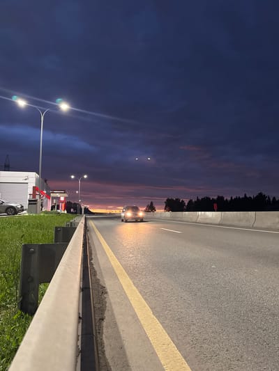 This vertical, low-angle, eye-level shot depicts a roadside scene at dusk, likely in or near the city of Можга, Russia. The foreground features a concrete barrier and a grassy verge with a metal guardrail. The asphalt road stretches into the distance, marked by a yellow line and a white line. A single car with its headlights on is driving on the road. In the mid-ground, a gas station with illuminated pumps and a building is visible on the left, with a parked car beside it. The sky is a deep twilight blue, transitioning to shades of pink and orange near the horizon, with some scattered clouds. Two streetlights with visible bulbs illuminate the scene. The background consists of a dark line of trees.