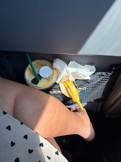 The image depicts the interior of a commercial transport vehicle, likely an aircraft, within the geographic context of Newark, United States. A person's bare upper right thigh and lower leg/foot are visible on the left side of the frame. The individual wears a light-colored garment patterned with black hearts.
Positioned on a dark grey textured floor or storage area, beneath a lighter grey panel, are several items. A clear plastic cup with a dome lid contains a light yellow, frothy beverage, possibly a smoothie or blended drink, topped with brown specks. A green plastic straw is inserted into the lid, which also bears a circular white sticker. To the right of the cup, several crumpled white paper napkins are present. A ripe, yellow banana peel with brown speckles rests on a black mesh seat-back pocket. Indistinct dark objects are partially visible within this mesh pocket. Sunlight casts strong shadows across the surfaces, indicating illumination from an external source, likely a window. No other individuals or specific identifying landmarks of Newark are present.