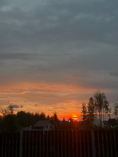 The image shows a sunset over a rural area in Gubakha, Russia. The sky is filled with dramatic clouds ranging from dark grey at the top to fiery orange and red near the horizon. The sun is setting, casting a warm glow. In the foreground, a dark wooden fence runs horizontally across the bottom of the frame. Behind the fence, there is a line of trees, some with lush green foliage and others appearing bare or thin. In the middle ground, several houses with pitched roofs are visible, nestled among the trees. One house appears to be under construction with exposed brickwork. Further in the distance, a blue-domed building, possibly a church, stands out against the setting sun. The overall scene depicts a peaceful twilight in a village setting. There are no people or animals visible in the image.