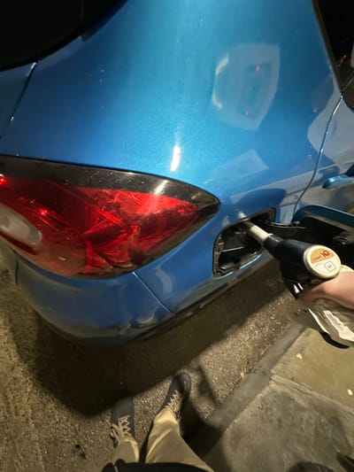 A shiny blue car being refueled with a black pump and a fuel nozzle. The car's taillight is bright red with intricate internal patterns. A hand holds the pump, partially obscured by a white cloth. The ground is wet and grainy, with a curb visible in the upper right. Two pairs of legs wearing light-colored pants and sneakers stand on the ground. The scene is captured at night, illuminated by artificial light.