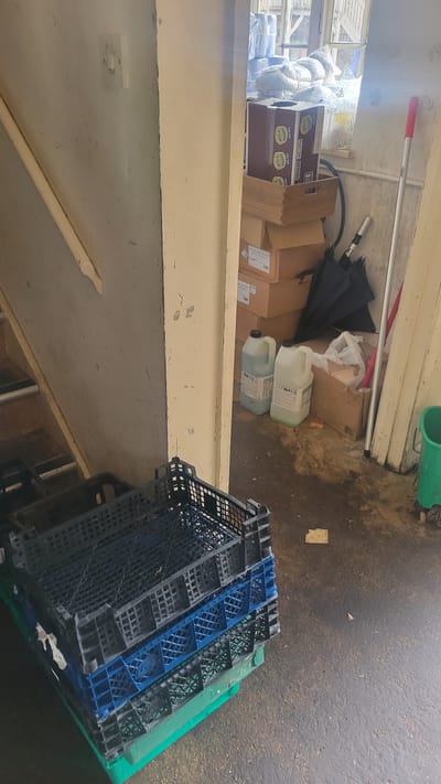 A cluttered interior scene, likely a storage area or back room, is shown in this image. In the foreground, several plastic crates are stacked, with a large black one on top, a blue one below it, and a green one at the bottom. To the right, in the background, are two large jugs of liquid, several cardboard boxes stacked, an umbrella, and a mop with a red handle. A bright green bin is partially visible on the far right. The floor appears to be concrete and shows signs of wear and some debris. The overall impression is one of disorganization and storage. The location is identified as Lacock, United Kingdom.