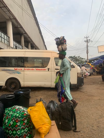 This is a medium shot, outdoor photograph taken during the day in Elelenwo, Nigeria. The scene depicts a man carrying a large, stacked load of bags and parcels on his head. He is wearing a light blue traditional outfit and a green cap. He is standing next to a white minibus with "Silver Travels Shuttle" written on its side. In the foreground, there are several bags and a black bucket on the ground. The background shows a building, electrical poles, wires, and other vehicles and market stalls. The ground is paved with rough asphalt. The sky is overcast.