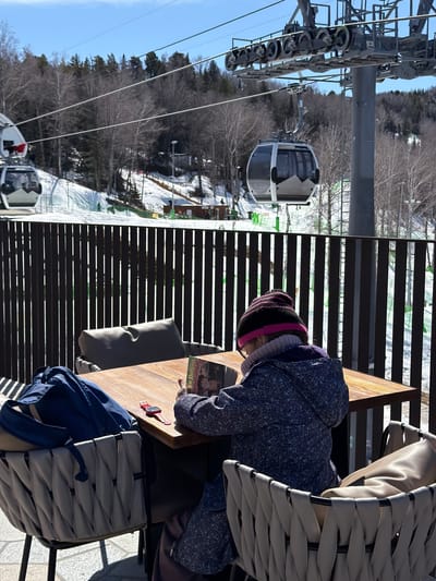 A person in a patterned blue jacket and black and pink hat sits at a wooden table in an outdoor setting. The person is reading a book. A red watch and a blue jacket sit on the table. The chair is made of woven material and appears to be on a patio. In the background is a ski lift and snowy terrain, suggesting a ski resort or mountain area in Ozernoe, Russia. A dark-colored railing separates the patio from the snow-covered landscape.