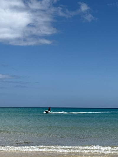 A bright, daytime image captures a serene coastal scene under a clear blue sky. The top two-thirds of the frame are dominated by the expanse of the sky, featuring scattered, wispy white clouds that are more concentrated towards the upper left. The bottom third of the image depicts calm ocean waters meeting a sandy shore.
In the ocean, a person is visible on a dark surfboard, appearing to be surfing or paddleboarding near the shoreline. The individual is partially obscured, wearing what appears to be a dark or red top. A thin line of white foamy water trails behind the surfboard, indicating movement. The water is a clear, light blue-green hue, with small, gentle waves breaking into white foam as they approach the sandy beach on the right side of the frame. The beach itself is a light, sandy strip.
The scene suggests leisure and outdoor activity on a sunny day. The weather is clear and bright, consistent with a pleasant beach environment. There is no visible text in the image.