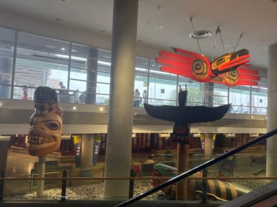 This image shows the interior of a modern building, likely an airport terminal, with large windows offering a view of the exterior. The scene is dominated by several large, intricately carved wooden totems. One totem, a tall, upright figure with an expressive face and bared teeth, stands prominently. Another, a large carved bird with spread wings, hangs suspended from the ceiling, painted in bold red and black colors. Additional totem pole pieces, some with vibrant patterns, are visible at a lower level.

Several people are visible in the background, seen through the windows and on different levels of the building, suggesting a public space. Some appear to be walking or waiting. The lighting suggests it's daytime, and the view through the windows shows an overcast sky. Blue banners with the text "YVR" are displayed on pillars, indicating the location is Vancouver International Airport (YVR) in Richmond, Canada. The overall atmosphere appears calm and functional, typical of an airport environment, with the art installations providing a cultural and visual focal point.