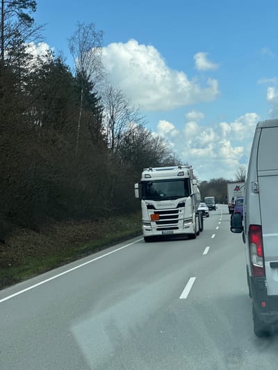 The image depicts a two-lane asphalt road, likely a highway or major rural route, under a bright blue sky with scattered cumulus clouds. The foreground shows the rear-right portion of a white cargo van, including a red vertical taillight assembly and a black side mirror.

In the opposing lane, a white Scania semi-trailer truck is approaching. Its front features a hazardous materials placard displaying "30" over "1202," indicating the transport of diesel fuel. The license plate "LA-XH 840" is visible on the truck's front. Behind the Scania, several other vehicles are visible, including a dark-colored bus or large van and a white passenger car.

The road features dashed white lane markings and a solid white line delineating the left edge of the carriageway. To the left of the road, a dense, wooded area is present, composed of a mix of bare deciduous trees, including a prominent tall birch, and dark green coniferous trees. The ground immediately adjacent to the road is dark earth with sparse green growth.

The setting is a roadway environment consistent with rural Bavaria, Germany. Based on the "LA" prefix on the Scania truck's license plate (Landshut district) and the requested location, the scene is situated on a road in or near Kirchdorf am Inn, Germany.