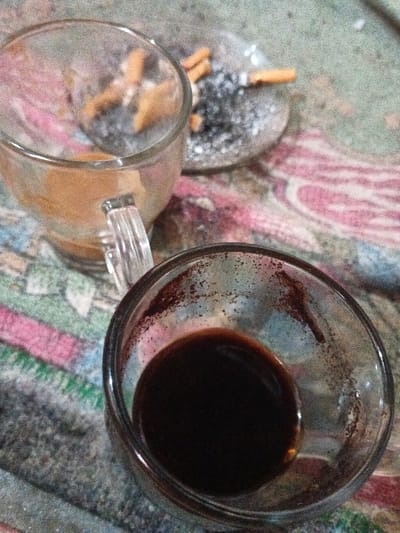 This image presents a top-down view of a table surface in Pyingyaung, Myanmar (Burma). In the foreground, a clear glass cup contains a small quantity of dark liquid residue and dark particulate matter adhering to its interior. Behind it and slightly to the left, a clear glass mug with a handle holds light brown residue at its bottom. Further in the background, a circular clear glass ashtray is visible, containing multiple extinguished cigarette butts, significant ash, and some general debris. One cigarette butt retains its filter and shows evidence of being partially smoked. All items are arranged on a patterned textile surface, likely a tablecloth or mat, featuring stripes and paisley-like designs in shades of green, pink, red, and blue. No individuals are present in the scene.