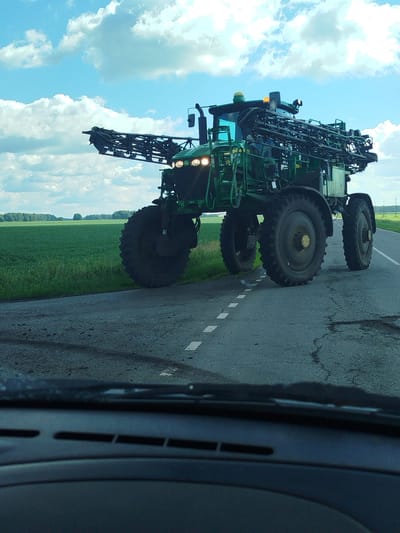 A large, green, self-propelled sprayer with extended booms is driving on a paved road. The sprayer has large tires and is equipped with lights on its top. The road has a dashed white line. To the left of the road is a vast, green field, and in the distance, a line of trees under a blue sky with white clouds. The image is taken from inside a vehicle, with the dashboard and windshield wipers visible in the foreground. The sprayer is positioned centrally in the frame, with its booms extending to the left. The overall scene depicts agricultural machinery in operation in a rural environment.