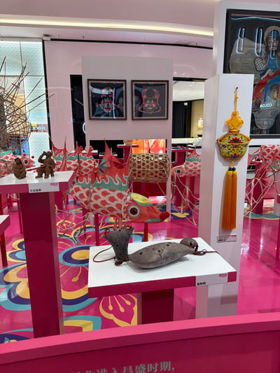 The image depicts an indoor exhibition space, likely a retail environment, with a vibrant pink floor adorned with a colorful, abstract floral pattern. Various display stands, also predominantly pink, are arranged throughout the scene, showcasing a collection of decorative items.

On a white display table in the foreground, two pottery figures are placed: one standing and one seated. Behind these figures, several red and white fish-shaped decorations with flame-like fins hang suspended. To the right, on another white display, is a decorative ceramic item with ornate yellow and blue patterns, featuring a prominent orange tassel. Below this, a textured grey, oblong object rests on a white surface.

Further into the background, two framed artworks are mounted on a white wall. These appear to be intricate depictions, possibly of traditional figures or scenes, with a dark frame. Another display features a woven basket. Additional fish-shaped decorations are visible in the mid-ground and background. The overall impression is one of a festive and visually engaging exhibition, likely for a cultural or seasonal event. The text on the display labels is in Chinese. Based on the Chinese characters visible on the display labels, the location is likely the city of Nanjing in Jianye District, China.