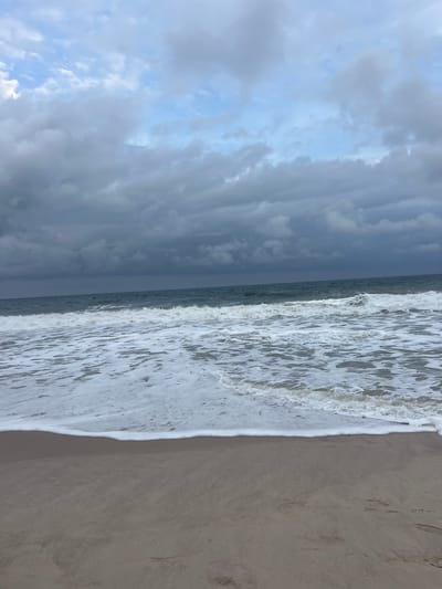 This media file captures a beach scene, likely in Mopo-Akinlade, Nigeria, with a focus on the meeting of the ocean and the shore. The image is oriented vertically, presenting a view from the sandy beach out towards the water and the sky.

The scene is dominated by natural elements: the sand of the beach, the ocean with its waves, and the sky filled with clouds. The sand appears to be a light tan color, with some subtle textures and footprints visible. The ocean is depicted with white foamy waves breaking gently onto the shore, creating a dynamic contrast with the calmer water further out. The water itself is a deep gray-blue, suggesting a slightly overcast or moody atmosphere.

The sky is a significant part of the composition, filled with a dramatic display of clouds. These clouds range in color from a bright, pale blue peeking through in some areas, to dark, heavy gray formations that suggest impending rain or a storm. The lighting suggests it could be late afternoon or early evening, with the diffused light from the overcast sky contributing to the muted tones of the scene.

There are no people or man-made objects visible in the frame, emphasizing the natural beauty and tranquility of the location. The overall emotion conveyed by the image is one of natural power and serenity, with the vastness of the ocean and the sky meeting the quiet expanse of the beach. No text is visible in the media file.