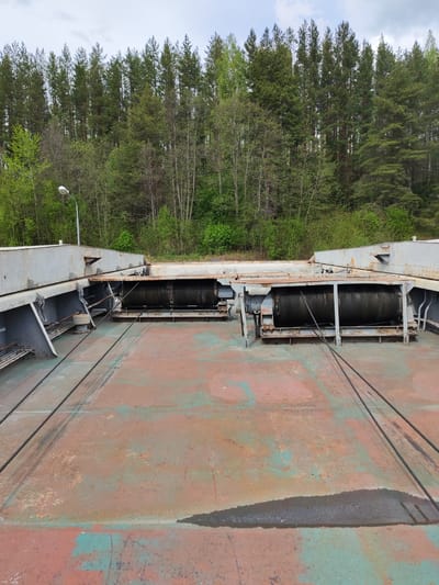 The image shows a section of what appears to be a dam or large industrial structure in Alekseevskoe, Russia. Two large, rusted metal winches with wound cables are visible in the center, anchored to a gray metal structure. The floor is a large expanse of rusted metal plating with patches of green and red paint. In the background, a dense forest of coniferous trees is seen, and a solitary street lamp extends from a structure on the left. The sky is overcast. No people are visible.