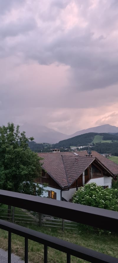 The image captures a twilight scene in Deutschnofen - Nova Ponente, Italy. In the foreground, a dark metal railing is visible, separating the viewer from a grassy area with a rustic wooden fence. Beyond the fence, a residential building with a red tiled roof dominates the midground. The building has white walls, wooden shutters, and illuminated windows, suggesting occupancy. A large, leafy green tree is positioned to the left of the building. In the background, rolling hills covered in dark green forests recede into the distance, culminating in a hazy mountain range under a soft, pinkish-purple sky filled with dramatic, cloud-laden twilight. A ski slope is visible as a lighter green swath on one of the distant hills. The overall atmosphere is serene and hints at the transition from day to night.