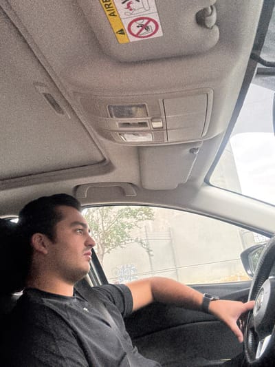 The image displays the interior of a vehicle, viewed from the front passenger seat, showing a male driver and the external environment in Santa Catarina, Mexico.
A male individual, with dark hair and short facial hair, is seated in the driver's position, oriented in a left-facing profile. He wears a black short-sleeved t-shirt. His left arm is bent, and his left hand, wearing a dark-colored smartwatch, rests on the steering wheel at approximately the 8 o'clock position.
The vehicle's interior features a light grey headliner with a visible sunroof opening and an overhead console housing lights and buttons. A light grey sun visor on the passenger side is partially visible, displaying a yellow "AIRBAG" warning label with pictorial warnings. The steering wheel is black with a central emblem.
Through the driver's side window, green foliage and a chain-link fence are visible. The view through the front passenger window shows a light-colored, possibly concrete or stucco, wall with some graffiti markings at the lower left, beneath an overcast sky. The scene suggests the vehicle is located in an urban or suburban area of Santa Catarina, Mexico.