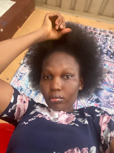 A woman with dark skin and afro-textured hair lies sideways on a patterned surface, looking directly at the camera. She wears a navy blue dress with pink and white floral patterns. Her arm is extended towards the upper left, with her hand slightly clenched. The background includes what appears to be a collage of images, possibly from magazines or posters. Natural light highlights her features, creating a casual and intimate atmosphere.