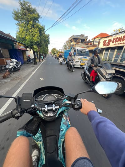 The image shows a first-person perspective from a motorcycle traveling on a street in Abiansemal, Indonesia. The street is paved and lined with shops and trees. In the foreground, the handlebars and dashboard of the motorcycle are visible, showing a speed of 26 km/h. The rider's hands and legs are also in view. Several other motorcycles and a truck are on the road, indicating moderate traffic. The sky is clear and blue. The shops on the left have closed shutters and advertisements. A dog is visible on the sidewalk. A sign for "KEMBE Bakery & Cake Sibok" is prominent on the right side of the street.
