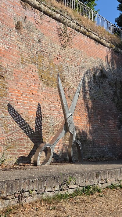 A pair of large, metallic scissors sculpture stands in front of a brick wall. The scissors are positioned with the blades open and facing upwards. The sculpture is located on a paved pathway, with a low brick wall running along the edge of the pathway in the foreground. Beyond the brick wall, dry grass and fallen leaves are visible. The brick wall behind the sculpture is weathered and has some patches of moss and vines growing on it. A white metal railing runs along the top of the brick wall, with some dry grass and greenery growing above it. The sky above is clear and blue. The overall scene suggests an outdoor public space. This sculpture is located in the city of Ulm, Germany.