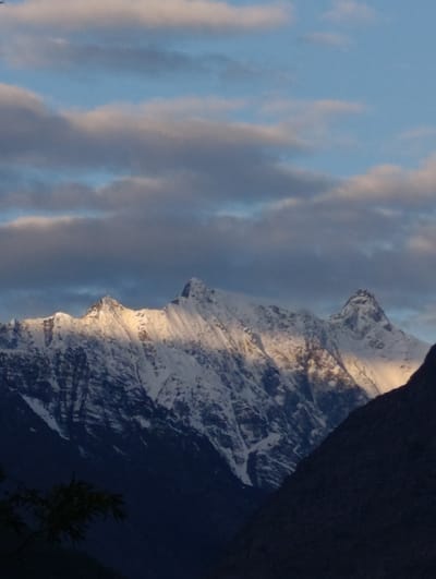 The image captures a majestic view of snow-capped mountains under a sky with scattered clouds. The sunlight illuminates the peaks, highlighting the rugged terrain and creating a dramatic contrast with the shadowed valleys. The foreground is dominated by dark, silhouetted slopes, with a hint of vegetation visible on the left. The scene suggests a remote, mountainous region, likely at a high altitude. The image does not contain any people or any indications of urban environments within the frame.
