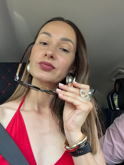 A woman is in the driver's seat of a car, holding a pair of sunglasses in her right hand. She is wearing a red halter top and large, ornate jewelry, including statement earrings, a chunky bracelet, and a ring on her right index finger. Her makeup is noticeable, with bold lipstick and defined eyebrows. The interior of the car is visible, with black upholstery and a seatbelt. The setting appears to be outdoors, possibly in a scenic area given the light conditions and hint of greenery in the background through the car window. The location is Chvizhepse, Russia.
