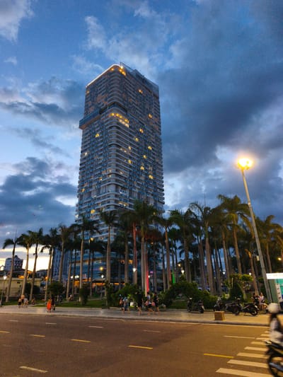 The image shows a tall, modern building with rounded balconies and illuminated windows, set against a cloudy, twilight sky. Palm trees line the foreground and flank the building, creating a tropical urban scene. A street with visible lane markings runs in front of the palm trees, and several people are walking along a paved area beside the street. There are also parked motorcycles and a streetlamp casting a warm glow. The building's architecture suggests it is a hotel or residential high-rise. The presence of palm trees and the warm ambient lighting indicate a warm climate. The sign on the right side of the image includes the text "AN PHU". This, along with the architecture and tropical vegetation, strongly suggests the location is a coastal city in Vietnam. Given the context of An Nhon city in Vietnam, this scene depicts a vibrant, contemporary urban environment characterized by modern architecture, lush tropical foliage, and pedestrian activity.