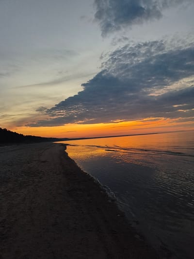 This is a photograph of a beach at sunset in Apšuciems, Latvia. The sky is a gradient of colors, from pale gray at the top to vibrant orange and yellow near the horizon. Dark, textured clouds are scattered across the upper portion of the sky. A thin line of dark trees forms the silhouette of the coastline on the left. The beach itself is composed of dark sand, and the edge of the water is visible on the right, reflecting the colors of the sunset. The water is calm, with subtle ripples. There are no people or man-made objects visible in the image. The scene is tranquil and natural.