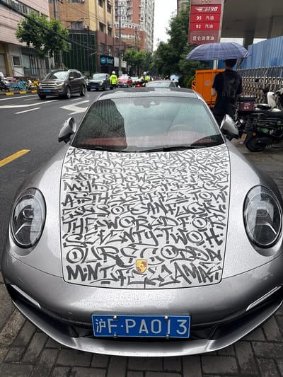 This urban street scene, likely in Jing'an District, China, is captured on an overcast day with recent rain, as indicated by visible water droplets on surfaces. The focal point is a silver Porsche sports car, possibly a 911, parked roadside. Its hood features a distinctive black-and-white graphic wrap, adorned with abstract, graffiti-style letters and shapes, with the iconic Porsche emblem still visible. The car's front displays a blue Chinese license plate, "沪F·PAO13".
To the left, a person, partially seen, holds a blue and white plaid umbrella, suggesting current or recent rainfall. Behind them stands a red sign detailing fuel options and prices in Chinese: "98 汽油", "95 汽油", "92 汽油" (98, 95, 92 Gasoline), and "昆仑润滑油" (Kunlun Lubricants). An orange utility cart is also visible in this area.
The background reveals a bustling city environment with multiple parked and moving vehicles, including cars, motorcycles, and bicycles. Buildings line the street, interspersed with trees. The ground consists of asphalt road with a yellow line and a brick sidewalk. The image portrays a lively urban setting, highlighted by the personalized sports car.