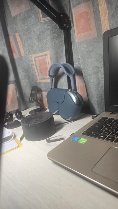 The image depicts a desk scene with various electronic devices and accessories. In the foreground, a laptop is partially visible, showing its keyboard and trackpad with stickers indicating Intel Pentium and NVIDIA GeForce graphics. To the left of the laptop, a pair of blue over-ear headphones is hanging from a black microphone stand. In front of the headphones, there is a cylindrical black speaker. Further to the left, a black phone stand is positioned behind a small black object that could be a camera lens cap or a similar accessory. A green pen rests on a yellow notepad. A small clear plastic object, possibly a USB drive or memory card case, is near the speaker. The background features a patterned curtain. There are no people visible in the image. The scene appears to be indoors, likely a home office or study. No specific landmarks or features identifiable with the city of Ukhta, Russia are present in the image.