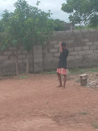 The image depicts a young person performing a plank exercise in an outdoor environment. The location is Achara Ndiagu, Nigeria. The individual, wearing a dark-colored shirt and red shorts, is positioned parallel to a concrete block wall. Their hands are placed flat on the ground in front of them, and their body is held in a straight line from head to heels. The ground is reddish-brown and appears to be a dirt surface. Vegetation, including trees and grass, is visible in the background. A concrete block is located nearby.