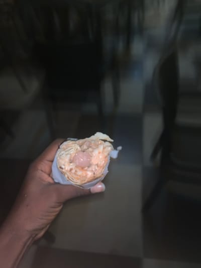 A dark-skinned hand holds a half-eaten wrap, presumably a shawarma, encased in white paper. The wrap's filling is visible, showing shredded cabbage, a creamy orange sauce, and a spherical pink meatball-like ingredient. The background is out of focus but shows what appears to be a kitchen setting with checkered wall tiles and stainless-steel appliances. The image is slightly dark and grainy.