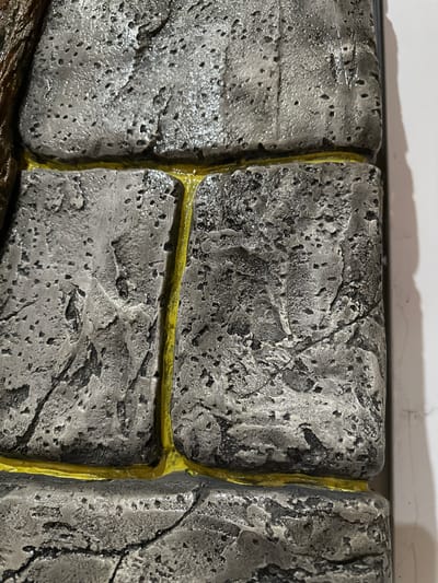 The image shows a close-up, angled view of a textured surface resembling aged, grey stone blocks with bright yellow grout. The stone has a rough, porous texture with visible pits and cracks, and appears to be wet or have a glossy finish in some areas. The yellow grout runs in thin, irregular lines between the blocks, highlighting their irregular shapes and placement. The lighting suggests a studio or controlled environment, casting soft shadows. There are no people, animals, or any other contextual elements visible in this image, and no information is provided about its location in Kalininskaya, Russia.