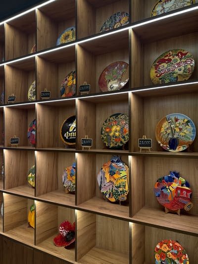 The image shows a shelf filled with decorative plates and fish-shaped ceramic art pieces, each displayed with a small price tag indicating a cost of 23,000 or 28,000 (currency unspecified). The shelves are made of light-colored wood, and the lighting is provided by linear LED strips running vertically between the shelves. The scene is situated in the city of Tsakhkadzor, Armenia, where such handcrafted art and ceramics are sold.