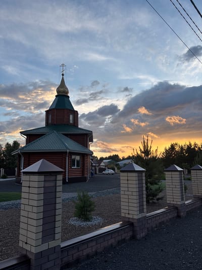This image shows a small, wooden church with a green, multi-tiered roof and a golden dome topped with a cross. The church is situated in a residential area, evidenced by the brick fence and houses in the background. The setting appears to be a rural or suburban location in Surak, Russia, given the presence of trees, gravel pathways, and a white car.
The time of day is sunset, with a dramatic sky filled with clouds illuminated by warm, orange and yellow hues. The sunlight casts a soft glow, suggesting a peaceful and serene atmosphere. The weather appears to be clear with scattered clouds, typical of an evening in this region. There are no people visible in the image, and the focus is on the architecture of the church and the natural beauty of the sunset. The overall mood is tranquil and picturesque.