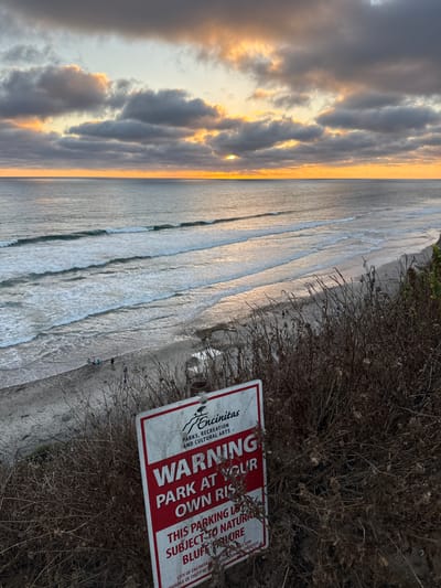 A sunset view over the ocean is captured from a cliff overlooking a beach in Encinitas, California, United States of America. A red and white warning sign is prominently displayed in the foreground, partially obscured by dry brush. The sign reads: "WARNING PARK AT YOUR OWN RISK. THIS PARKING LOT IS SUBJECT TO NATURAL BLUFF FAILURE. CITY OF ENCINITAS IS NOT RESPONSIBLE FOR DAMAGE OF THEFT TO VEHICLES OR CONTENTS." The ocean exhibits gentle waves, and a few individuals are visible on the sandy beach. The sky is filled with dramatic clouds tinged with orange and yellow from the setting sun.