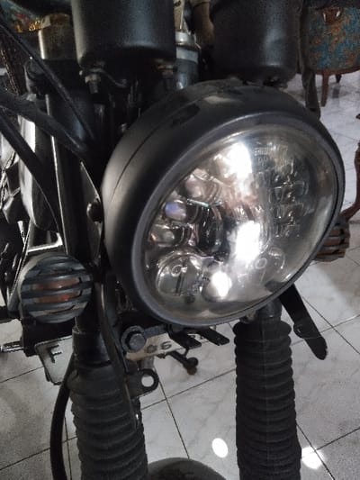 The image displays the front section of a dark-colored motorcycle or similar two-wheeled vehicle, observed indoors on a tiled floor in what is stated to be Majdalena, Czechia. No individuals are present.

The central subject is a circular LED headlight, featuring a clear lens revealing multiple internal emitters and reflectors, encased in a matte black housing. It is mounted to the front forks. To the immediate left of the main headlight, a smaller, circular auxiliary light with a horizontal slatted grille covering an amber or orange-toned lens is visible. Black rubber gaiters (bellows) enclose the front suspension forks on both sides. Various metallic mounting brackets, bolts, and sections of wiring are visible. A small white rectangular object, possibly a license plate, displaying the letter "F," is affixed near the left fork assembly.

The floor beneath the vehicle consists of light-colored, reflective square tiles with dark grout lines, showing reflections from an overhead light source. In the blurred background to the upper right, a distressed, possibly rusted, blue-painted object with visible legs is partially discernible, suggesting a cluttered indoor environment consistent with a garage or workshop.