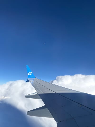 The image shows an airplane wing against a backdrop of blue sky and clouds. The airplane wing is grey, and a blue vertical stabilizer can be seen on the wing's edge, featuring a white logo. White clouds border the wing, contrasting with the clear blue sky. In the distance, a small, light-colored object is visible in the sky. The image was taken from inside an airplane. The location in San Fernando de Henares, Spain is not discernible from the image.