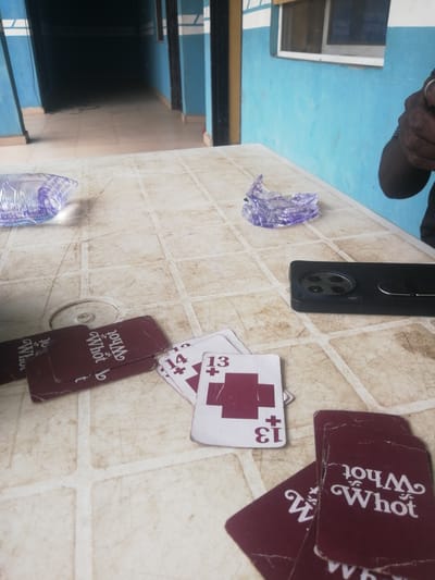 In Ajasse Ipo, Nigeria, a scene unfolds at a table, likely outdoors given the natural light and worn surface. On the table are several playing cards, some red with white text that reads "Whot", and others white with red markings and numbers such as "13" and "14". A black smartphone lies face down to the right of the cards. To the left, a partially consumed bottle of water sits on the table. In the background, a blue-painted corridor stretches into darkness, suggesting an interior hallway. The overall setting appears casual and possibly implies a game in progress.