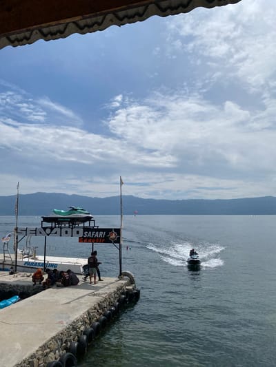 A jet ski is being ridden on a body of water near a pier in Tuk Tuk, Indonesia. The pier is made of concrete and lined with tires. A boat is docked at the pier, with a jet ski on a display stand above it. Several people are sitting on the pier, and one person is standing. A sign above the boat reads "SEA-DOO SAFARI SAMOSIR". The sky is cloudy, and mountains are visible in the background.