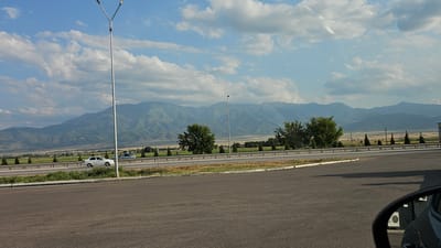 The image captures a wide landscape view of a highway with mountains in the background, likely situated near Жамбыл ауылдық округі, Kazakhstan. In the foreground, a paved area, possibly a parking lot or roadside, is visible, with a portion of a car's side mirror and body in the lower right corner. A white sedan is driving on the highway, which is bordered by green fields and a line of trees. Several tall streetlights stand along the highway. The sky is partly cloudy with dramatic formations of white and grey clouds against a blue backdrop. The mountains in the distance are layered and appear green and brown, with some areas showing shadow and light, suggesting a sunny day with some cloud cover.