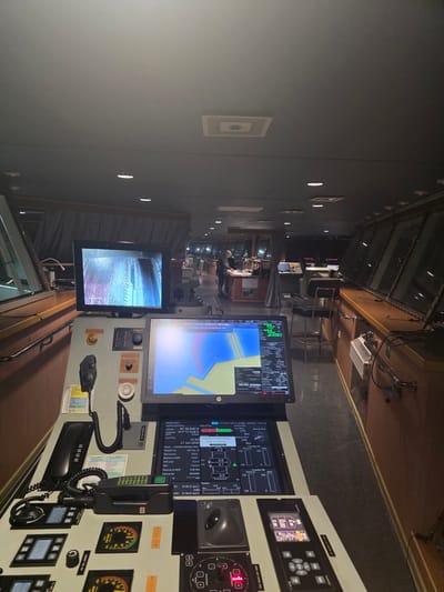 The image depicts the interior of a ship's bridge, likely a vessel docked or navigating near Brindisi, Italy, given the on-screen text referencing "CATANIA BRINDISI SH..." in the navigation display. The scene is dimly lit, with multiple screens displaying navigation and operational data. In the foreground, a complex control console is visible, featuring telephones, communication devices, and various buttons and dials. Two prominent monitors dominate the console: one displays a camera feed labeled "Camera 2 AFT RAMP," showing a textured, possibly metallic surface, and another shows a detailed navigational chart with overlaid data, including latitude, longitude, and speed. The chart indicates a location with coordinates 40°38.838' N and 017°57.628' E. Text on this screen also reads "System in SILENT Mode. Audible alerts switched off." Further back, another person is partially visible standing at a console, and a chair is positioned at a workstation. The bridge appears to be a functional command center with an expansive view through large windows, suggesting an operational or standby status for the vessel.