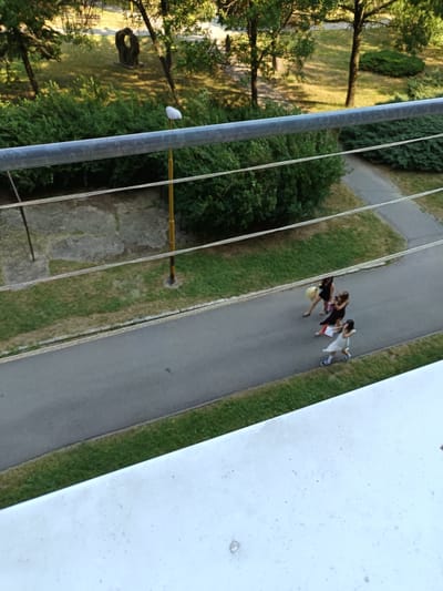 An overhead, slightly angled shot captures a paved pathway with a grassy verge on either side. Three young females are walking along the pathway from right to left. The one in front is wearing a white dress and the one in the middle is wearing a dark dress. The third, furthest away, is wearing a dark dress with a yellow object, possibly a balloon, to her left. The path is bordered by dense green bushes and trees with dappled sunlight on the ground. A metal railing runs horizontally across the top of the frame, with four thin wires stretched beneath it. In the background, a solitary yellow lamppost with a white light fixture is visible, along with a concrete slab and a dark, abstract sculpture. The foreground is dominated by a white, textured surface, likely a balcony or ledge. The scene appears to be an outdoor urban setting in the city of Košice, Slovakia.