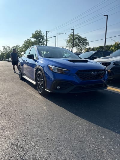 The image depicts a vibrant blue 2022+ generation Subaru WRX, parked diagonally in an asphalt lot under a clear, bright blue sky. The car is positioned with its front end facing slightly towards the right side of the frame. It features black multi-spoke wheels and black fender flares, with a distinctive red "WRX" badge on its front grille. To the left of the car, a man wearing a dark t-shirt, dark shorts, and white athletic shoes stands with his hands in his pockets, looking towards the left side of the frame, away from the camera.
In the background, a row of other vehicles, predominantly dark-colored SUVs, are parked in an orderly fashion, suggesting a car dealership lot. Partially visible to the right of the blue WRX is the front section of a white Subaru SUV. The ground is an asphalt surface with visible cracks and speckles, and strong sunlight casts distinct shadows across the pavement. In the upper background, mature green trees, tall utility poles with multiple overhead power lines, and streetlights are visible against the cloudless sky.
Based on the presence of multiple Subaru vehicles, including a new model WRX, and the typical environment of a new car sales lot, the location is identified as the parking lot of **Patrick Subaru, located at 1680 N Meacham Rd, Schaumburg, Illinois, United States of America**.