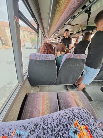 The image captures the interior of a public transport vehicle, likely a bus or train, from a tilted, elevated perspective looking down a narrow aisle. The vehicle features rows of seats with black main surfaces and patterned purple fabric on the sides of the seatbacks, adorned with colorful abstract designs in orange, red, blue, and black.

Multiple passengers are visible. In the foreground, a person with long reddish-brown hair is seated, facing away from the camera. Further down the aisle, a man with a beard, wearing a black t-shirt and light blue shorts, stands. Another man in a black shirt is seen looking towards the camera. Additional passengers are seated or standing in the background.

Large windows on one side of the vehicle provide an external view of buildings with tiled roofs and a white van, suggesting an urban or semi-urban setting. The bright lighting indicates daytime. A red sticker with partially legible white text, appearing to be a safety warning, is visible on the window frame. The scene captures everyday travel within a public transport setting, with the overall ambiance being functional and unadorned.