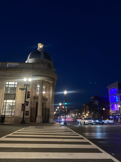 At night, a crosswalk is illuminated by streetlights and traffic signals on a city street. A bank, PNC BANK, with a dome and columns, is on the left side of the image. Cars are driving on the street, and a few people are visible in the distance on the sidewalk. The sky is dark blue and clear. The location appears to be in Washington, D.C. due to the street sign that reads "Wisconsin Ave".