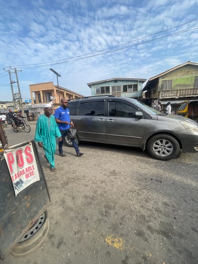 A medium shot captures an outdoor street scene in Akowonjo, Nigeria, under a partly cloudy sky. In the foreground, a black metal bin with a sign reading "POS IS AVAILABLE HERE" is positioned on the left. Two men are walking on the asphalt road. The man on the left is older, wearing a traditional turquoise robe and a white cap, and is walking to the left. The man on the right is younger, dressed in a blue polo shirt, dark jeans, and sunglasses, carrying a black handbag in his right hand. He is walking slightly behind and to the right of the older man. A grey minivan is parked on the right side of the frame. Behind the minivan, two men are conversing, and a yellow motorized rickshaw is visible. In the background, buildings with various architectural styles and colors line the street, along with a utility pole and electrical wires. Further in the distance, more buildings and a billboard are visible. A motorcycle with two occupants is seen on the left side of the image. The overall impression is a bustling urban environment.