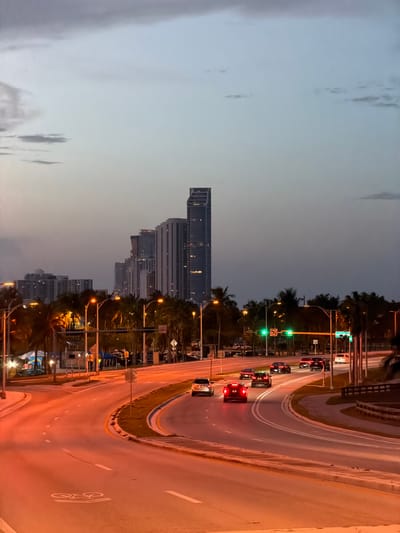 The image depicts a nighttime view of a multi-lane road in Bal Harbour Village, United States of America, with a city skyline in the background. The sky is a gradient of pale blue to dark gray, with some clouds visible. The road is illuminated by streetlights and the headlights and taillights of several vehicles. Tall buildings, characteristic of a city skyline, are visible in the distance, their lights glowing. Palm trees line the side of the road, adding a tropical element to the scene. The road curves through the foreground, with lane markings clearly visible. A bicycle symbol is painted on the road surface in the lower left portion of the image. Several cars are in motion on the road. Traffic lights are visible, with some showing green. The overall impression is that of an urban environment at dusk or early night.