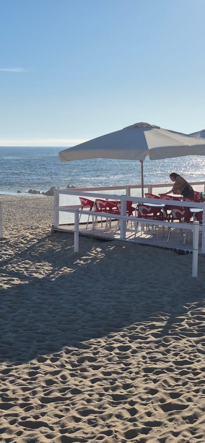 The image shows an outdoor seating area on a sandy beach, with the ocean in the background. A white umbrella provides shade over a series of tables and red chairs. A person, appearing to be a server, is visible behind the tables, possibly attending to them or serving customers. The beach is characterized by rippled sand, with shadows cast by the seating structure. The ocean is calm, with some rocks visible near the shoreline. The sky is clear and blue. This scene is likely part of a restaurant or cafe located on one of Vila do Conde's beaches, such as Praia da Ladeira or Praia do Forno.