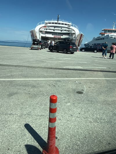 In Thassos, Greece, on a sunny day, a ferry named "AMIRA" is docked at a port, ready to embark vehicles. The ferry is large and white with red accents. In front of the ferry, several cars are lined up on the concrete pier, waiting to board. One car is on a trailer, carrying what appears to be a motorcycle or ATV. A man in a white shirt and dark pants stands near one of the cars, possibly a port worker. Further to the right, another ferry is docked, its upper decks visible. Two people, a man in a blue shirt and shorts and a woman in a patterned top and dark pants, stand observing the scene. In the foreground, a red and white striped traffic cone is positioned on the ground, casting a long shadow. The sky is a clear blue, and the sea is calm.