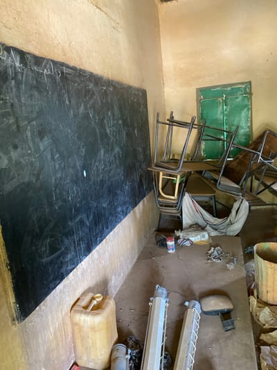 The image depicts a classroom in Khartoum, Sudan (15.552, 32.57). The classroom contains a large, black chalkboard with visible chalk markings, and a stack of overturned wooden and metal desks with their chairs on top. There is a green window in the back wall, and a bare, dirt floor. There is a plastic yellow jerrycan, a white cloth, a rusty metal toolbox, and a round metal object laying on the floor. In the background, a bucket can be seen. The room appears to be abandoned and in disarray.