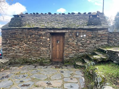 The image displays a small, single-story building located in Campillo De Ranas, Spain, constructed entirely of irregular, horizontally stacked dark grey and brown slate-like stones. The gabled roof is covered with flat slate tiles, partially obscured by green moss, and features a line of larger, rounded stones along its peak, with two distinct stone chimney-like structures. A central wooden door, comprising vertical planks, is equipped with a dark metal handle and a small, rectangular window with an internal grid in its upper half. To the right of the door, a dark metal wall lantern is affixed to the stone wall, positioned above two small, rectangular white utility plates. The ground immediately in front of the building is paved with large, irregular grey stone slabs, appearing wet in sections and interspaced with green weeds and scattered fallen leaves. To the right, an elevated area covered in green grass features rough stone steps leading upwards. A light-colored vehicle is partially visible on the far left. The upper portion of the scene shows a bright blue sky with scattered white clouds, alongside bare tree branches and distant foliage. No individuals or active subjects are present.
