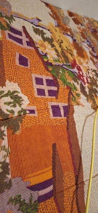The image displays a close-up, angled view of a woven textile, likely a tapestry or blanket, against a light background. The textile depicts a stylized house rendered primarily in orange hues, featuring purple-framed windows with white sills; one visible window has a four-pane grid, and another has two panes. Surrounding the house are woven representations of trees with autumn foliage, showing colors such as yellow, orange, green, and brown. A light beige or off-white background is also visible in the upper right. The weaving technique results in a textured, somewhat pixelated appearance of the depicted elements. A yellow electrical cable or cord runs vertically along the right side of the image, partially overlapping the textile. The scene is located in Maple Ridge, Canada.